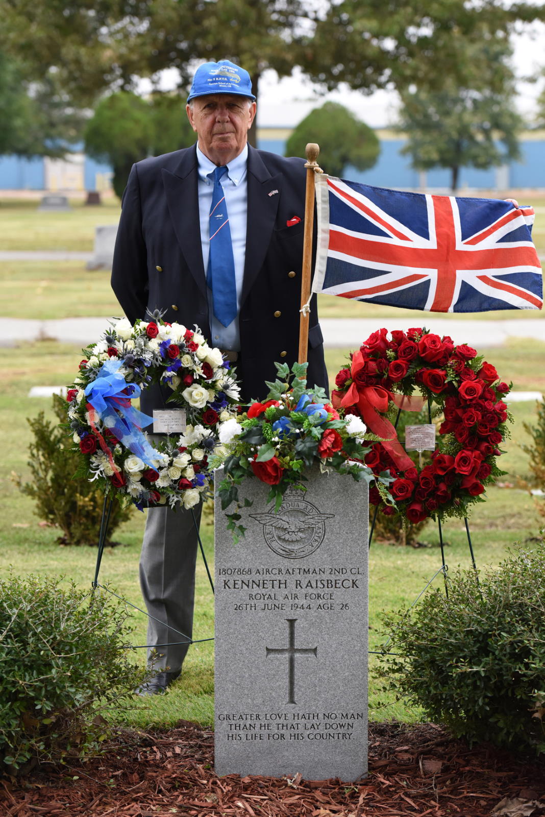 John Raisbeck son of British Cadet Kenneth Raisbeck standing next to his fathers grave marker with floral wreaths and flag of England